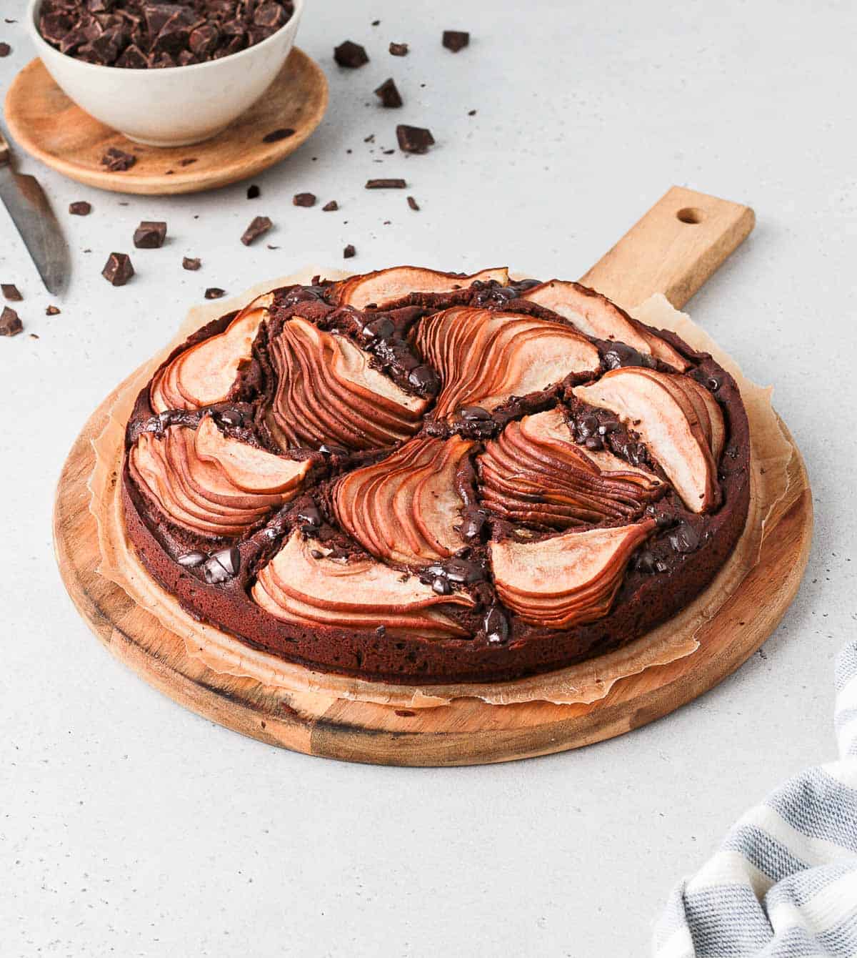 Chocolate pear cake on a round wooden board with chunks of chocolate.