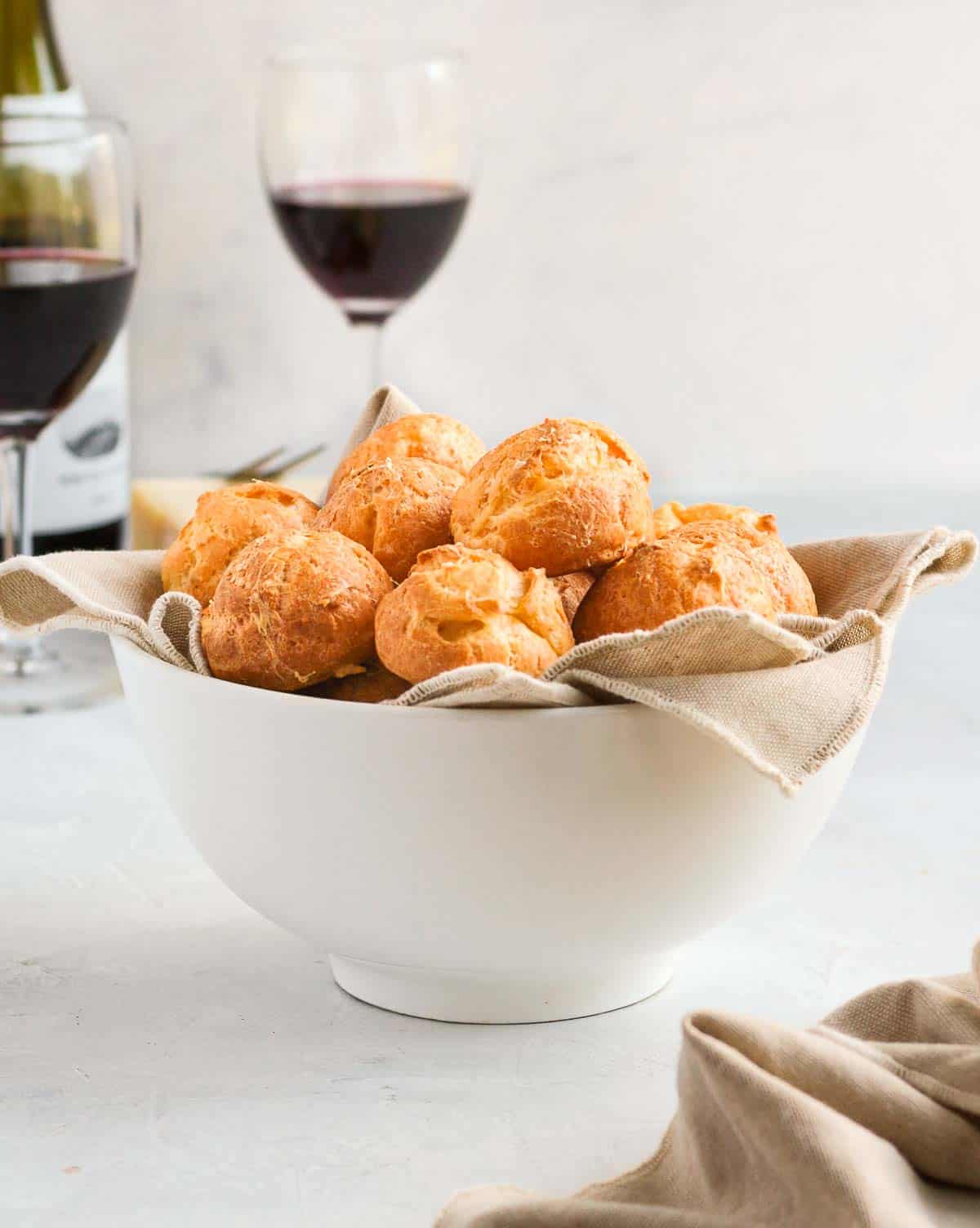 Gougeres in a white bowl with wine glasses in the background.