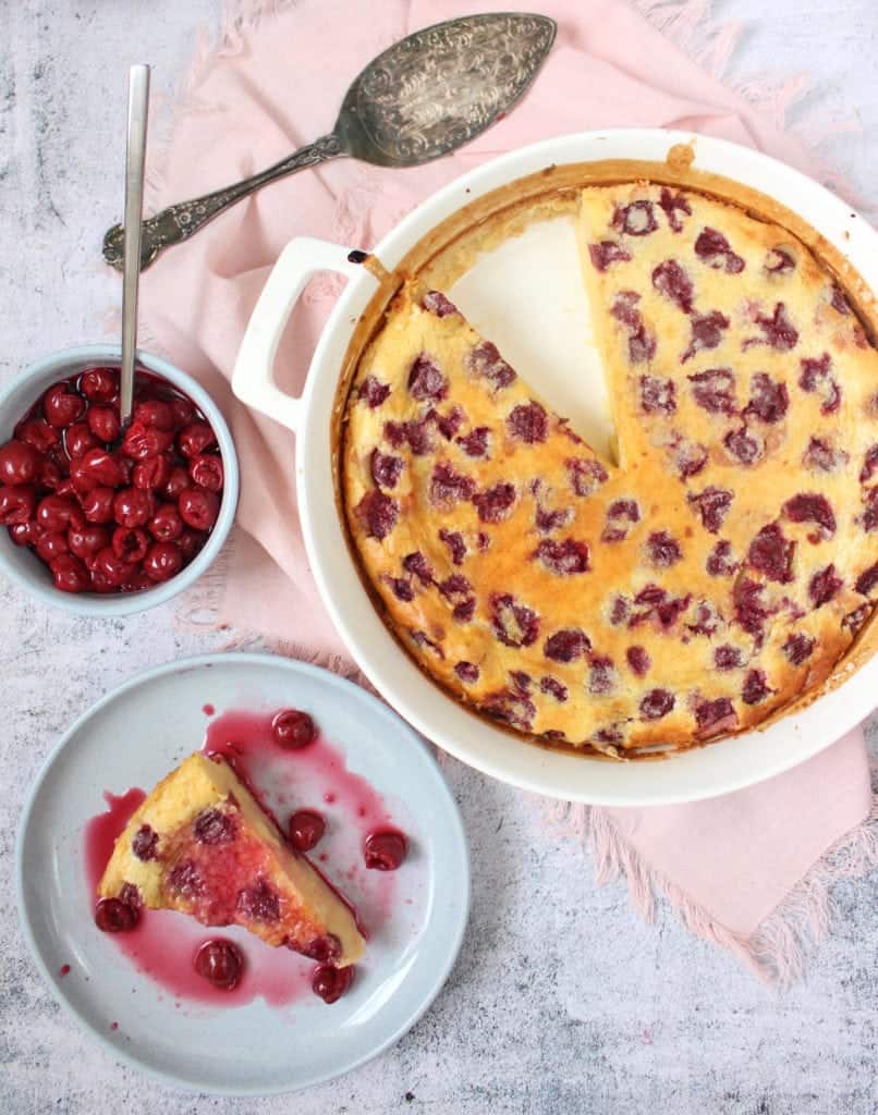 One slice of clafoutis on a blue plate next to the baking dish with the rest of the dessert and a bowl of morello cherries