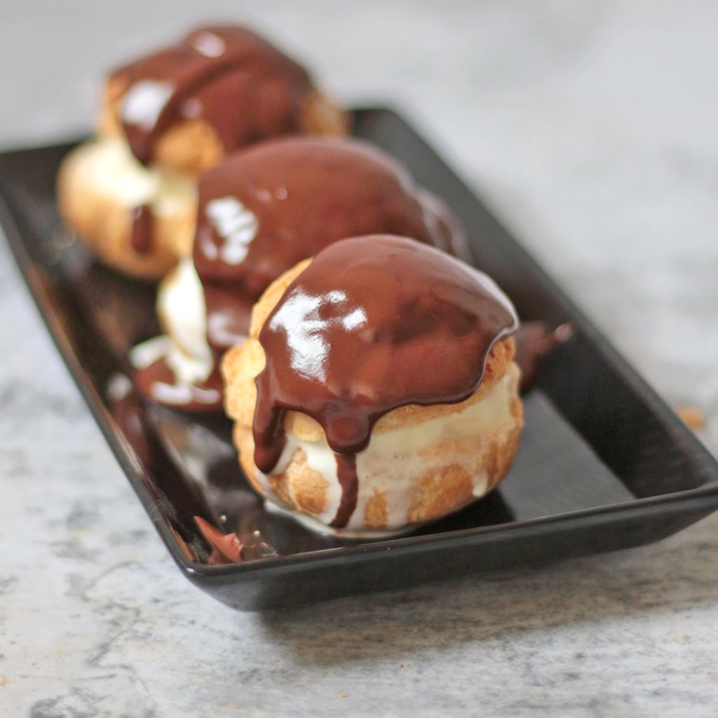 3 profiteroles with chocolate sauce on a black plate.