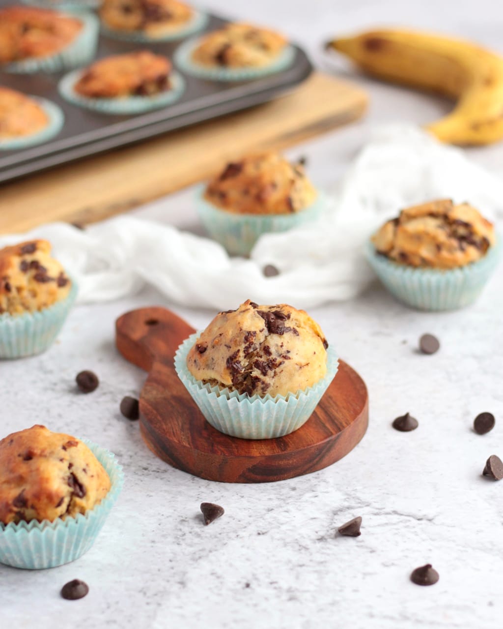 One muffin on small wooden board surrounded by muffins and chocolate chips