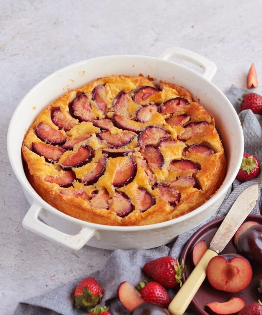 Clafoutis with Plums in the baking dish, surrounded by fresh plums and strawberries