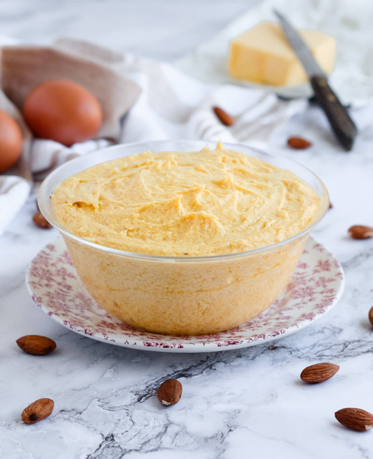 Cream in a glass bowl surrounded by almonds, eggs and butter.