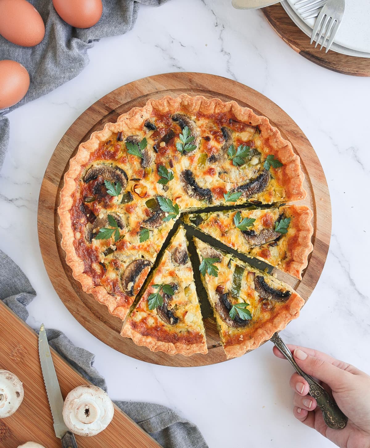 Quiche from above on a round wooden board with 3 slices cut and a hand picking up one slice