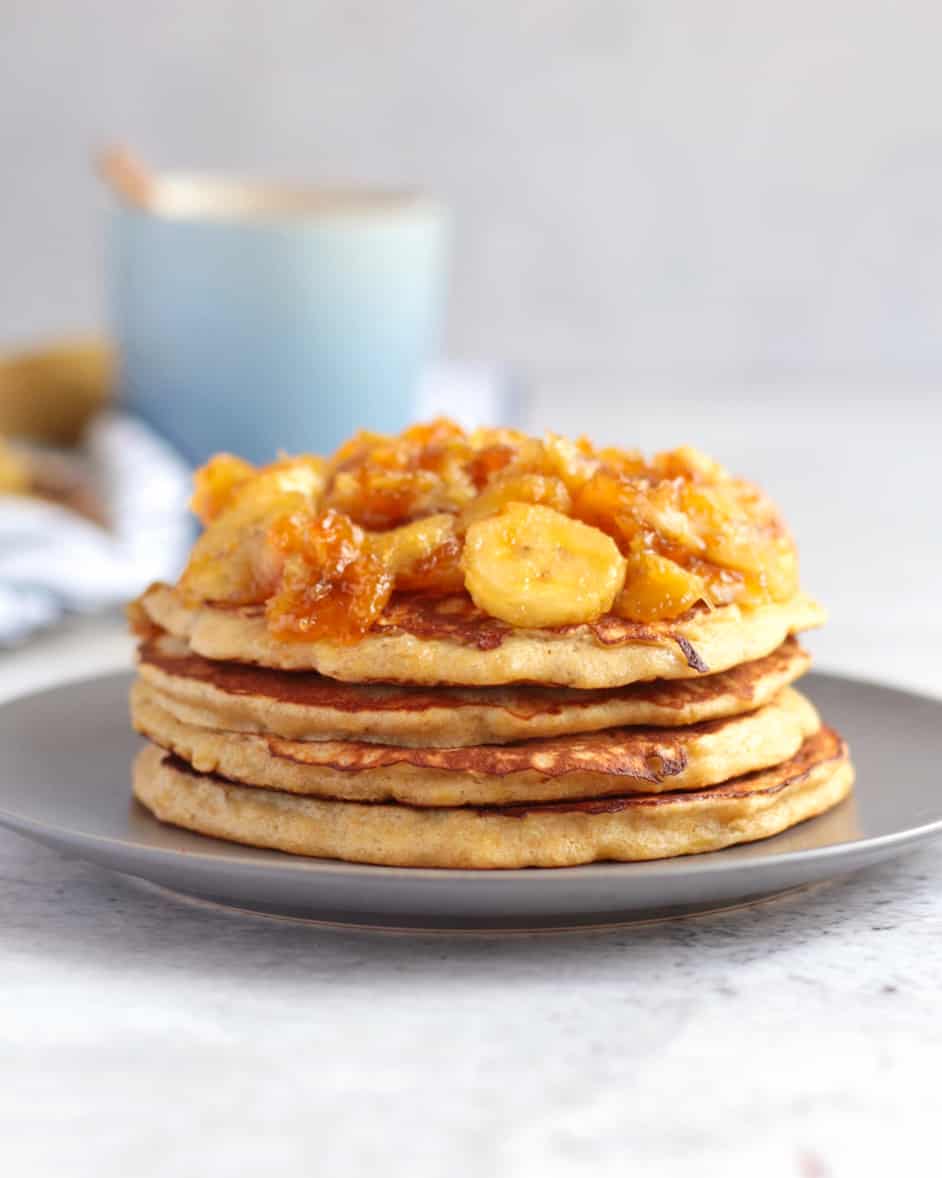 Stack of pancake topped with banana topping on a grey plate behind a white background.