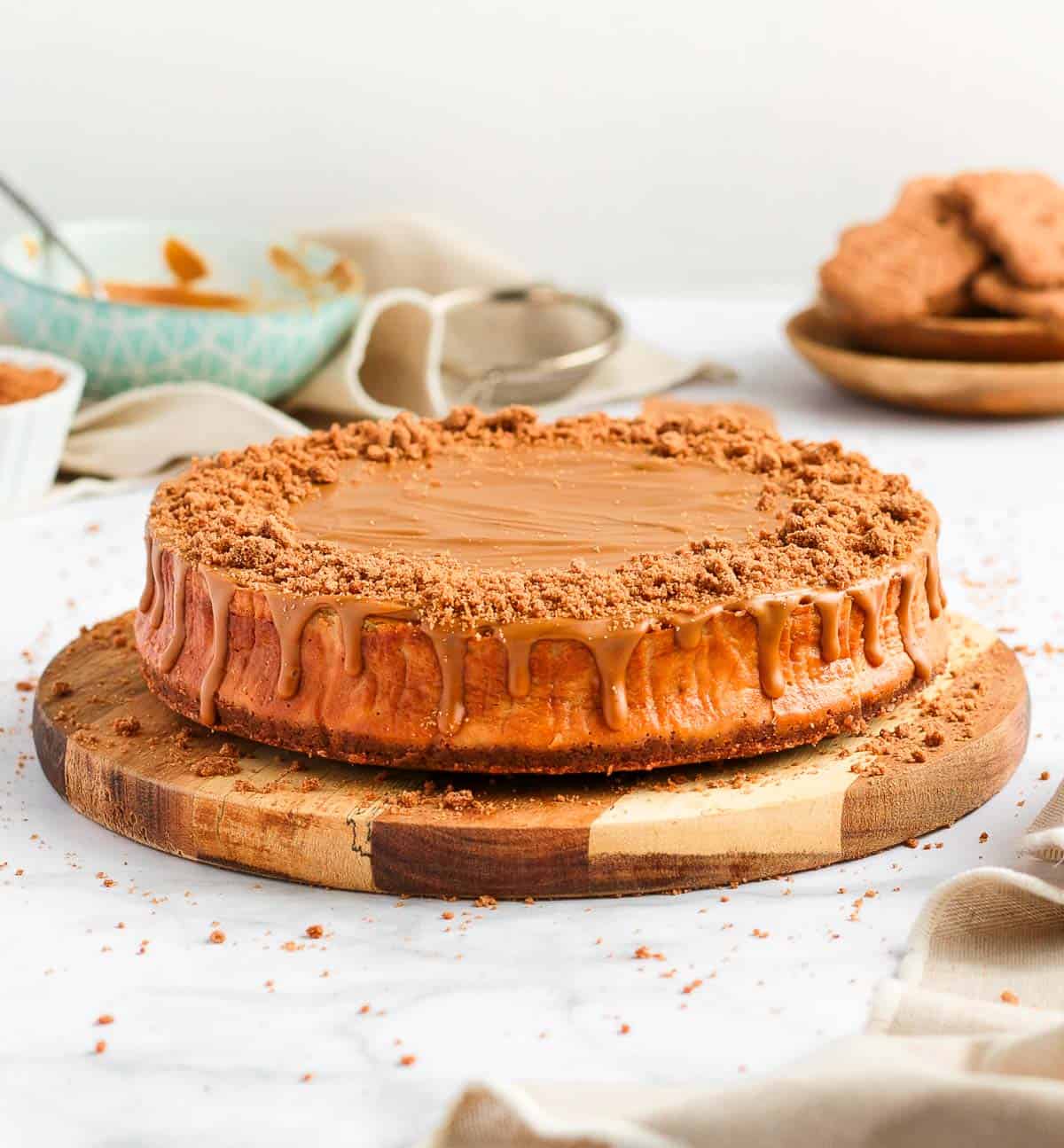 Biscoff Cheesecake on a round wooden board surrounded by speculoos cookies and a bowl with biscoff spread.