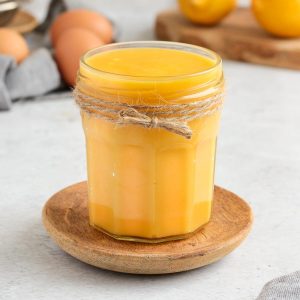 Lemon Curd in a glass jar over a small wooden plate.