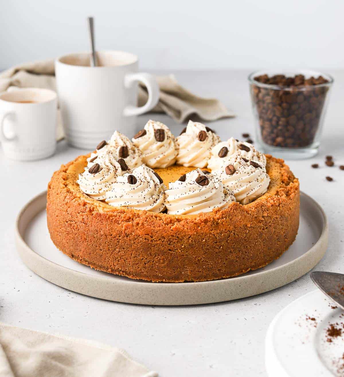 Cheesecake on a plate surrounded by coffee beans and cups of coffee.