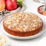 Cake on a large white serving plate surrounded by red apples.