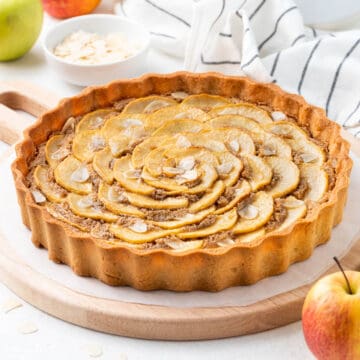 Apple tart on a wooden board surrounded by apples and a stripped tea towel.