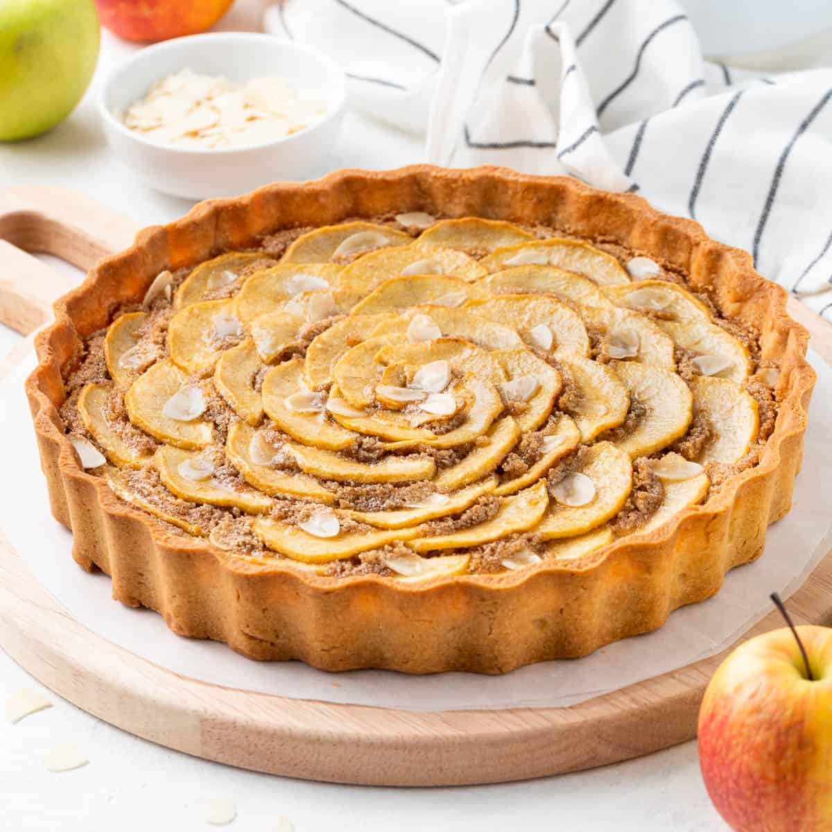 Apple tart on a wooden board surrounded by apples and a stripped tea towel.