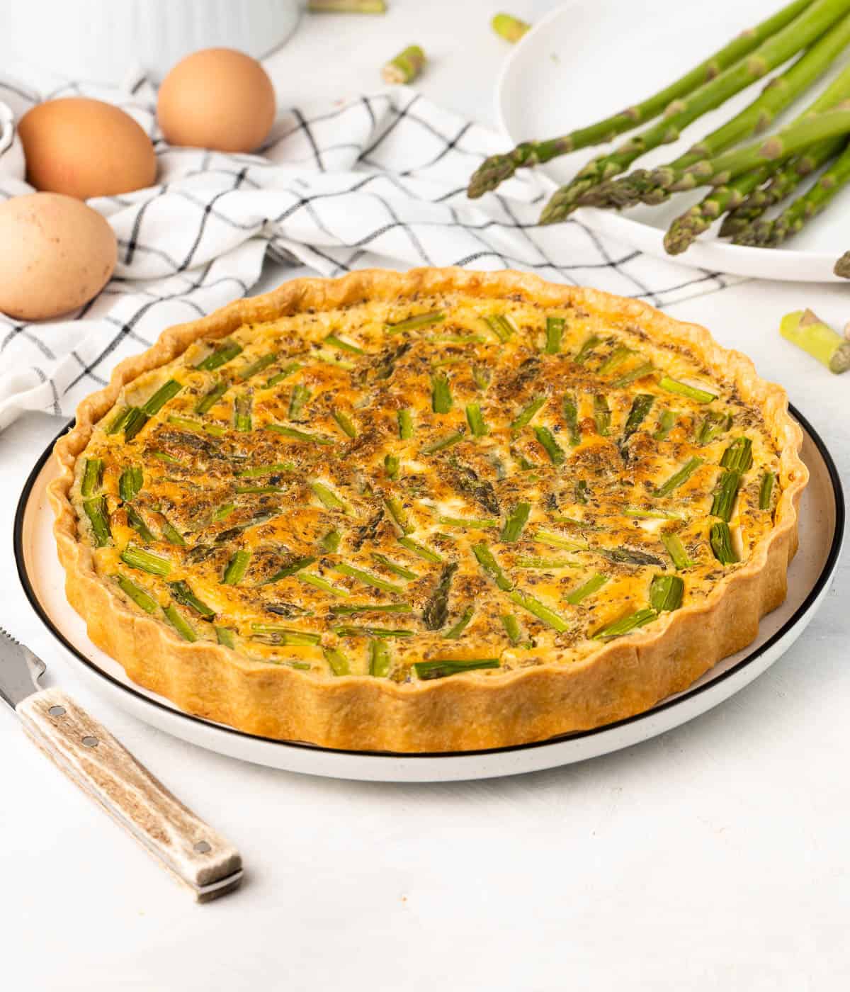 Side view on the quiche placed in a large white plate with ingredients in the background.