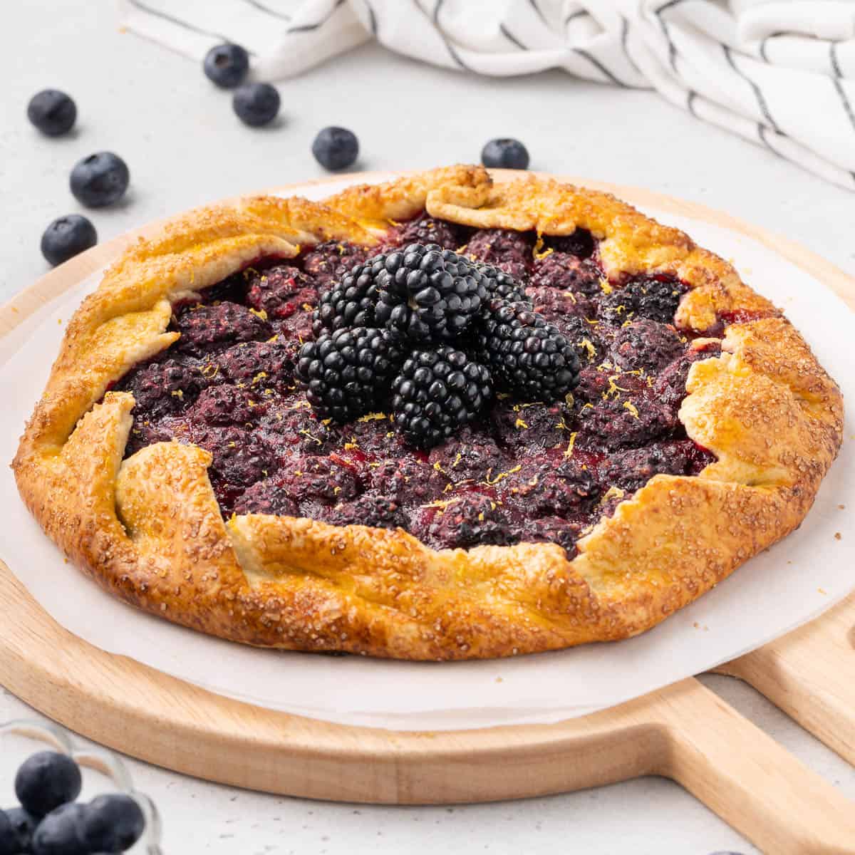 Tart topped with fresh blackberries on a sheet of baking paper over a round wooden board.