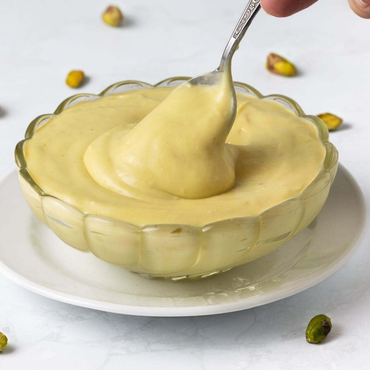 Pistachio cream dripping from a small spoon inside a glass bowl.