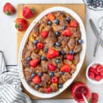 French toast baked in a white oval dish seen from above, placed on a rectangular cutting board and topped with fresh berries.