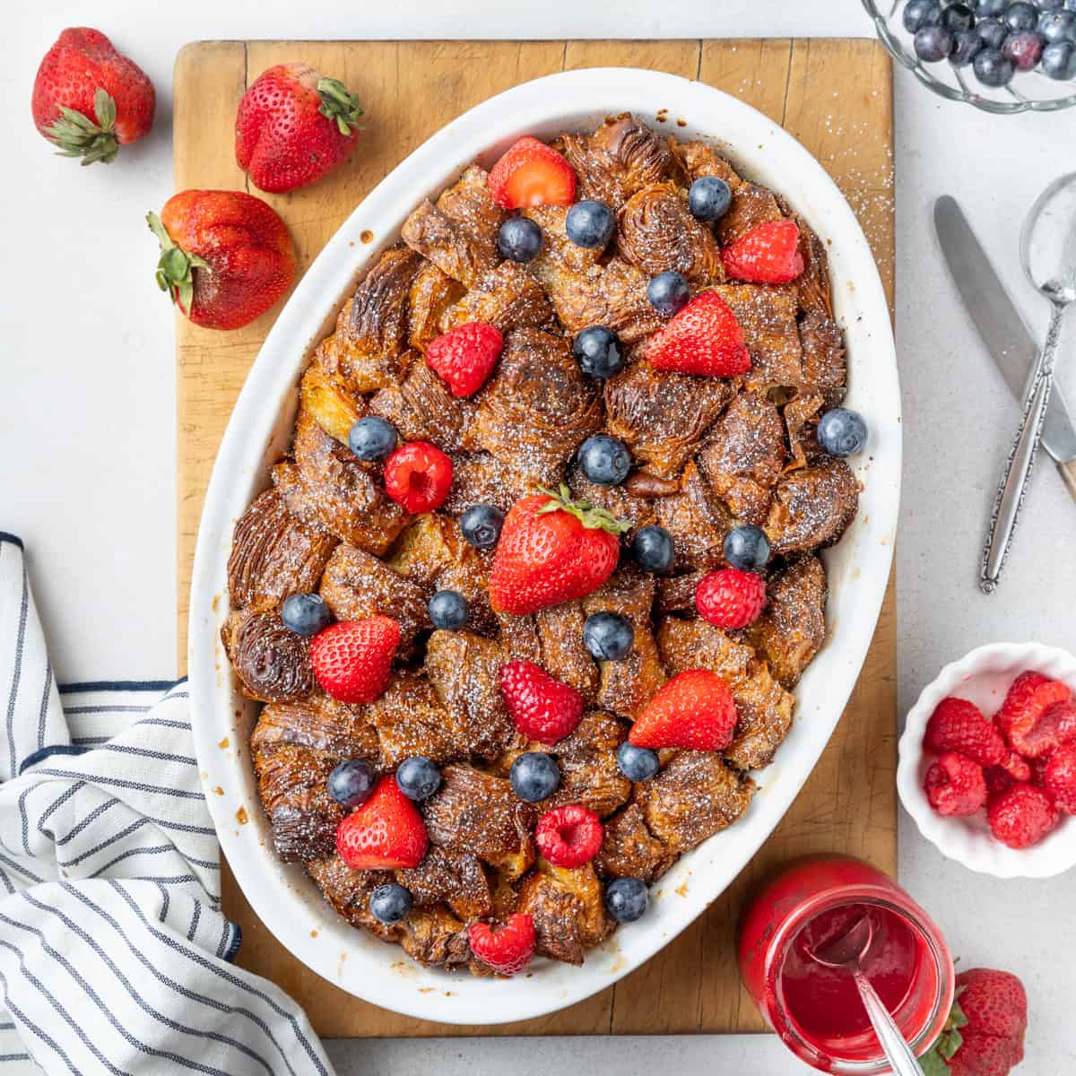 French toast baked in a white oval dish seen from above, placed on a rectangular cutting board and topped with fresh berries.