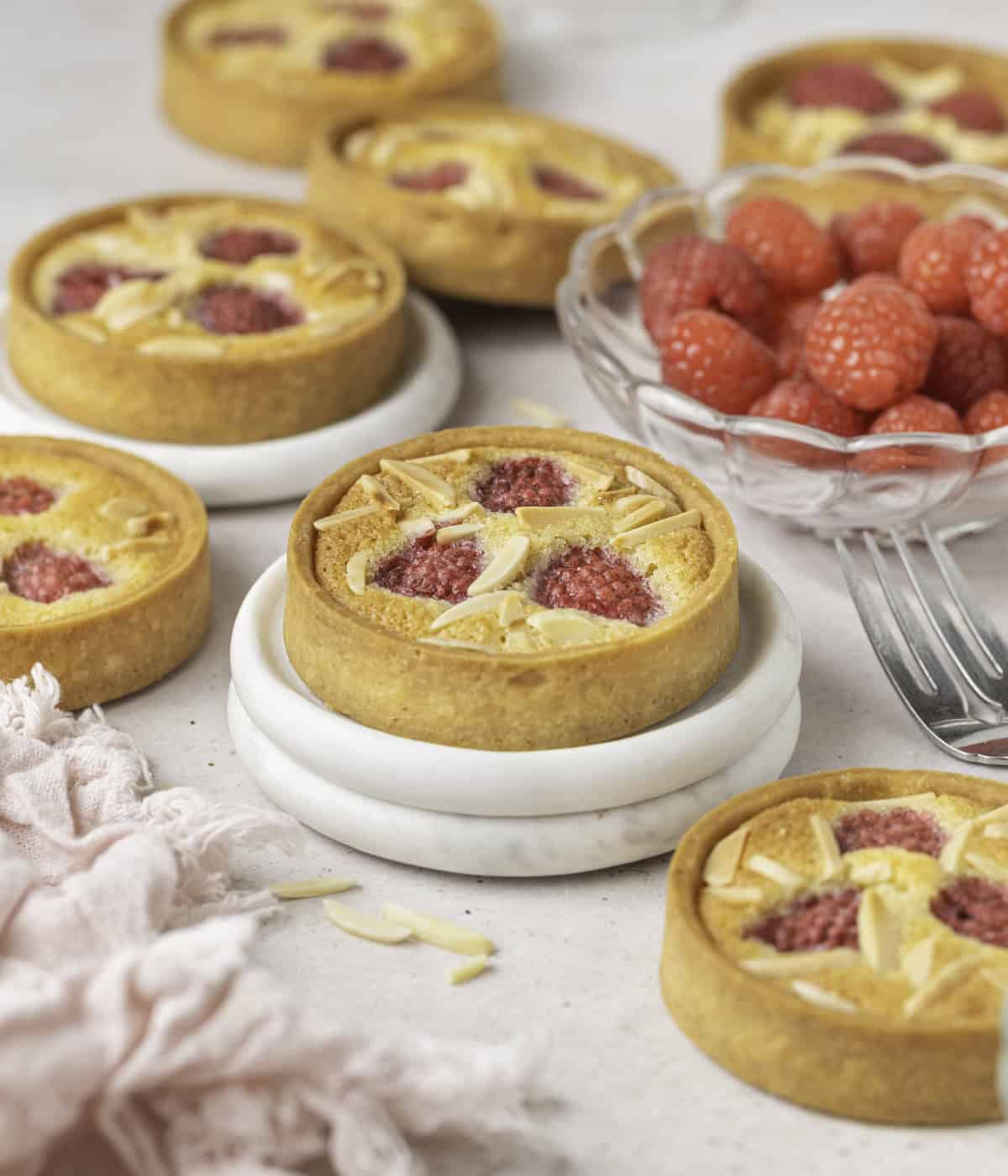 Tarts laid over a light beige surface with a light pink napkin and a bowl filled with fresh raspberries.